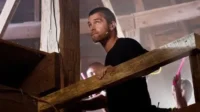 Banshee – T01E02:A Rave 8 Um homem sério com fone e camisa preta olha para cima de uma estrutura de madeira. No fundo,pessoas seguram luzes coloridas.