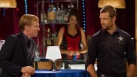 Banshee – T01E03:Conheça o Novo Chefe 7 Um homem de terno conversa intensamente com um xerife sorridente em um bar. Uma bartender serve ao fundo. O ambiente é acolhedor.