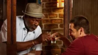 Banshee – T01E04:Meio Surdo é Melhor que Totalmente Morto 6 Dois homens em um bar brindam com shots. Um,mais velho de chapéu,olha o copo;o mais jovem sorri. Clima íntimo.