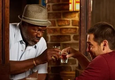 Dois homens em um bar brindam com shots. Um, mais velho de chapéu, olha o copo; o mais jovem sorri. Clima íntimo.
