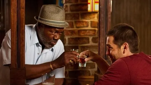 Dois homens em um bar brindam com shots. Um,mais velho de chapéu,olha o copo;o mais jovem sorri. Clima íntimo.
