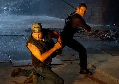 Cena de ação chuvosa. Homem de bandana ajoelhado, enfrentando outro de preto com bastão, que sorri. Ambiente escuro e molhado.