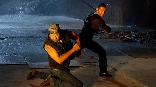 Cena de ação chuvosa. Homem de bandana ajoelhado,enfrentando outro de preto com bastão,que sorri. Ambiente escuro e molhado.