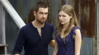 Banshee – T01E08:Viveremos Para Sempre 3 Homem com barba e mulher de vestido florido,ambos com expressões preocupadas,em cenário rústico com madeira e metal. Atmosfera sombria.