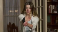 Banshee – T01E09:Sempre o Caubói 2 Mulher de cabelos claros,ferida e com sangue nas mãos,aponta uma pistola. Ambiente doméstico,clima tenso e perigoso.