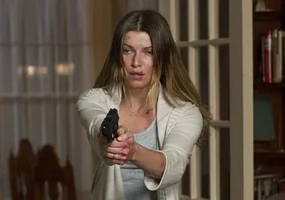 Mulher de cabelos claros, ferida e com sangue nas mãos, aponta uma pistola. Ambiente doméstico, clima tenso e perigoso.