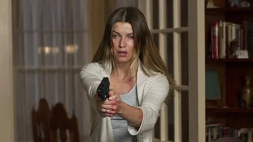 Mulher de cabelos claros,ferida e com sangue nas mãos,aponta uma pistola. Ambiente doméstico,clima tenso e perigoso.