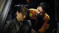 Mulher de uniforme futurista, olhos fechados, sentada em posto de comando escuro. Homem de laranja a observa com preocupação.