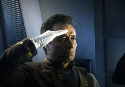 Homem de uniforme militar futurista escuro e luva branca saluda. Expressão séria em ambiente sombrio.