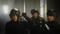 Mulher de capacete e dois homens em equipamento tático olham com intensidade em ambiente escuro. Clima de ação e suspense.