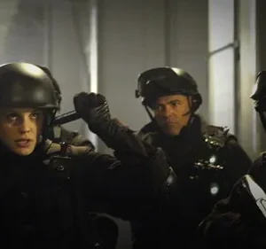 Mulher de capacete e dois homens em equipamento tático olham com intensidade em ambiente escuro. Clima de ação e suspense.