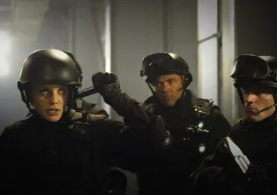 Mulher de capacete e dois homens em equipamento tático olham com intensidade em ambiente escuro. Clima de ação e suspense.