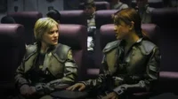 Duas mulheres em uniformes militares futuristas conversam em assentos de nave espacial. Uma loira, outra morena, ambas sérias.