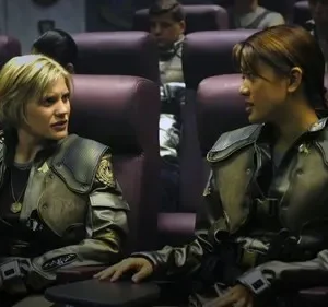 Duas mulheres em uniformes militares futuristas conversam em assentos de nave espacial. Uma loira, outra morena, ambas sérias.
