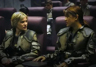 Duas mulheres em uniformes militares futuristas conversam em assentos de nave espacial. Uma loira, outra morena, ambas sérias.