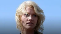 Mulher loira de cabelo ondulado olhando para cima com expressão séria, contra um fundo azul claro. Luz solar destaca o rosto.