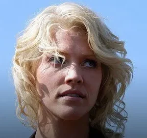 Mulher loira de cabelo ondulado olhando para cima com expressão séria, contra um fundo azul claro. Luz solar destaca o rosto.