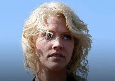 Mulher loira de cabelo ondulado olhando para cima com expressão séria, contra um fundo azul claro. Luz solar destaca o rosto.