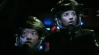 Dois personagens em capacetes de ficção científica olham fixamente, apreensivos, em uma cabine escura com luzes coloridas.