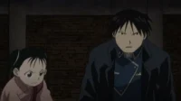 Roy Mustang e May Chang de Fullmetal Alchemist têm expressões sérias,olhando para frente,contra uma parede escura de tijolos.