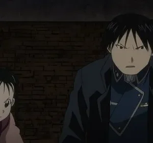 Roy Mustang e May Chang de Fullmetal Alchemist têm expressões sérias, olhando para frente, contra uma parede escura de tijolos.