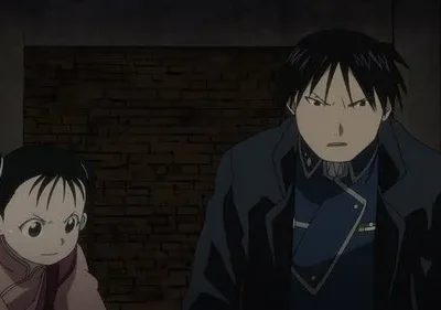Roy Mustang e May Chang de Fullmetal Alchemist têm expressões sérias, olhando para frente, contra uma parede escura de tijolos.