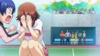 Em Grand Blue, Chisa, envergonhada, esconde o rosto enquanto Iori grita. Outros personagens e público assistem num campo esportivo.
