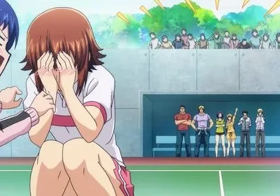 Em Grand Blue, Chisa, envergonhada, esconde o rosto enquanto Iori grita. Outros personagens e público assistem num campo esportivo.