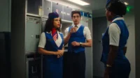 Três comissários de bordo em uniformes azuis e brancos estilo retrô conversam na galley de um avião. Uma mulher tem um lenço vermelho.
