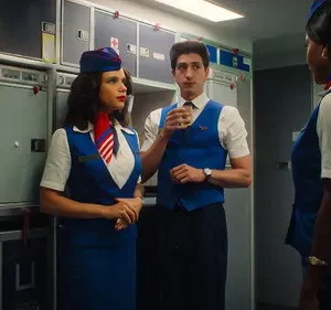 Três comissários de bordo em uniformes azuis e brancos estilo retrô conversam na galley de um avião. Uma mulher tem um lenço vermelho.