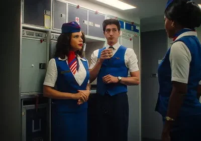 Três comissários de bordo em uniformes azuis e brancos estilo retrô conversam na galley de um avião. Uma mulher tem um lenço vermelho.