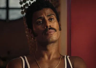 Homem negro de bigode, brinco e regata, com olhar sério em ambiente escuro e parede avermelhada ao fundo.
