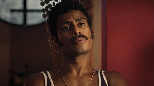 Máscaras de Oxigênio Não Cairão Automaticamente – T01E02:Canto de Oxóssi 1 Homem negro de bigode,brinco e regata,com olhar sério em ambiente escuro e parede avermelhada ao fundo.