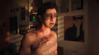 Jovem de torso nu, cabelos cacheados e olhos marejados, segura um cigarro. Quarto escuro com quadro de gato de óculos. Cena angustiante.