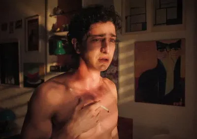 Jovem de torso nu, cabelos cacheados e olhos marejados, segura um cigarro. Quarto escuro com quadro de gato de óculos. Cena angustiante.