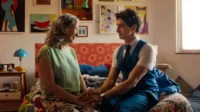 Mulher idosa e homem jovem de mãos dadas numa cama colorida. Eles sorriem em um quarto vibrante com arte nas paredes.
