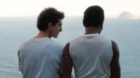 Dois homens na costa. Um olha para o perfil do outro, que observa o oceano calmo e uma montanha distante. Clima íntimo e reflexivo.