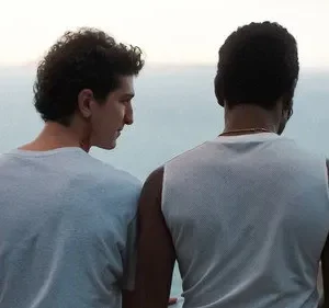 Dois homens na costa. Um olha para o perfil do outro, que observa o oceano calmo e uma montanha distante. Clima íntimo e reflexivo.