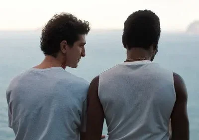 Dois homens na costa. Um olha para o perfil do outro, que observa o oceano calmo e uma montanha distante. Clima íntimo e reflexivo.
