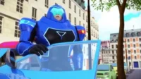 Homem em armadura azul futurista e surpreso, dirigindo carro azul com luzes vermelhas. Rua de cidade com prédios claros.
