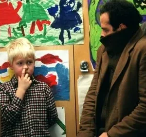 Monk: Adrian Monk, de casaco marrom, observa um menino loiro coçando o nariz. Fundo com arte infantil colorida. Cena peculiar e cômica.