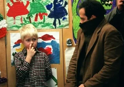 Monk: Adrian Monk, de casaco marrom, observa um menino loiro coçando o nariz. Fundo com arte infantil colorida. Cena peculiar e cômica.