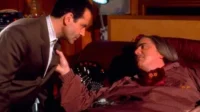 Adrian Monk de terno marrom inclina-se sobre um homem de cabelo comprido com almofada DB, reclinado no sofá. Clima sério em cena de Monk.