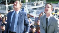 Em Office Space, dois homens de terno, um com bigode, olham curiosos para o alto em um evento ao ar livre. (108 caracteres)