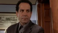 Adrian Monk (Tony Shalhoub), de terno escuro, com expressão séria e pensativa em ambiente interno. Imagem da série 'Monk'.