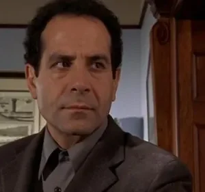 Adrian Monk (Tony Shalhoub), de terno escuro, com expressão séria e pensativa em ambiente interno. Imagem da série 'Monk'.