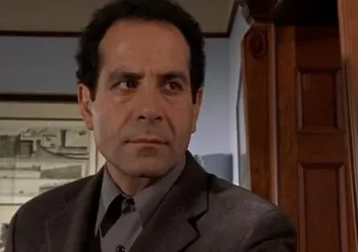 Adrian Monk (Tony Shalhoub), de terno escuro, com expressão séria e pensativa em ambiente interno. Imagem da série 'Monk'.
