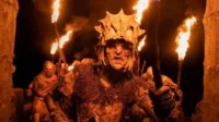 Um Orc de The Rings of Power com elmo e armadura lidera outros em um túnel escuro, iluminado por tochas. Atmosfera ameaçadora.