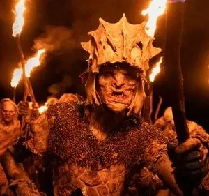 Um Orc de The Rings of Power com elmo e armadura lidera outros em um túnel escuro, iluminado por tochas. Atmosfera ameaçadora.