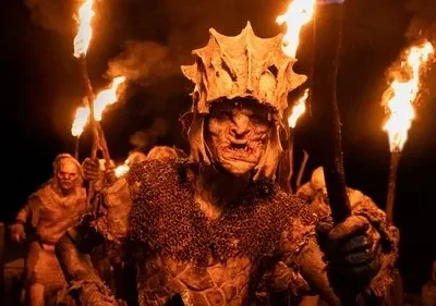 Um Orc de The Rings of Power com elmo e armadura lidera outros em um túnel escuro, iluminado por tochas. Atmosfera ameaçadora.